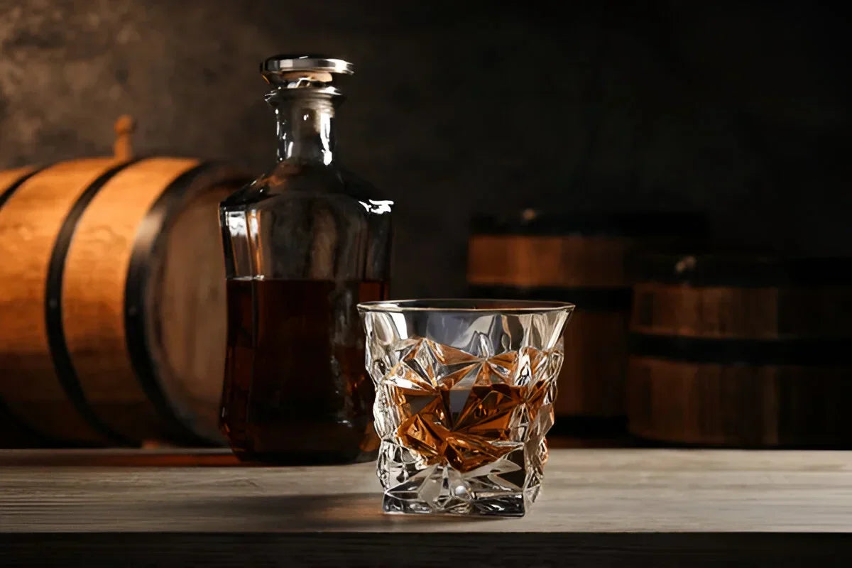 Verre à whisky taillé rempli de whisky ambré, posé devant une carafe et des fûts en bois de chêne dans une distillerie