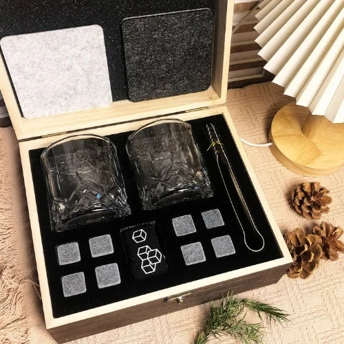 Coffret whisky ouvert avec verres, pierres réfrigérantes et sous-verres, accompagné de pommes de pin et décorations naturelles sur une table chaleureuse.