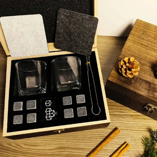 Coffret whisky élégant avec accessoires complets, présenté sur une table en bois avec pommes de pin et bâtons de cannelle en arrière-plan.