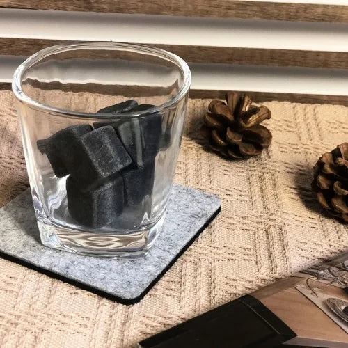 Verre à whisky transparent avec pierres réfrigérantes noires, posé sur un sous-verre en feutre, avec décorations naturelles en arrière-plan.