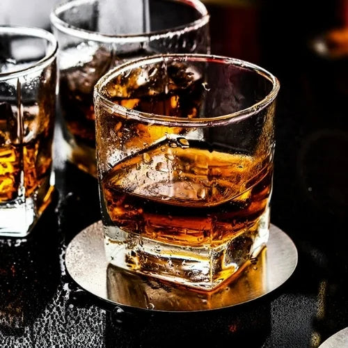 Un verre carré rempli de whisky, posé sur un sous-verre, brillant dans une ambiance festive et élégante.