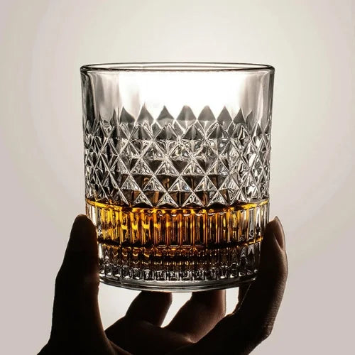 Verre à whisky en cristal texturé, rempli de whisky ambré, tenu à la main avec une lumière douce en arrière-plan pour mettre en valeur les détails du verre.