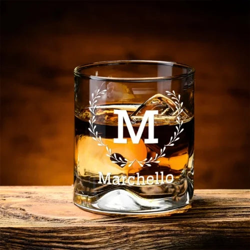 Verre à whisky personnalisé 'Marchello', posé sur une table en bois rustique, rempli de whisky ambré et de glaçons.