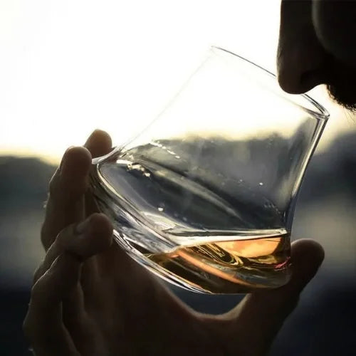 Homme savourant un whisky dans un verre de dégustation design, en contre-jour avec vue floue sur l'extérieur.