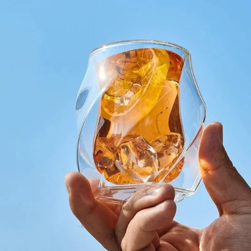 Verre à whisky au design original, rempli de whisky avec glaçons, tenu à la main sous un ciel bleu clair