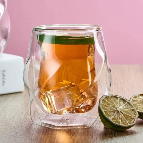 Verre à whisky au style unique, rempli de whisky avec glaçons et tranche de citron vert, posé sur une table avec fond rose.